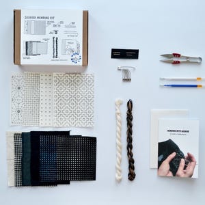 Può includere: Un kit di riparazione Sashiko con vari materiali. Il kit include una scatola con istruzioni, campioni di tessuto, aghi, filo, forbici e un libretto intitolato "Mending with Sashiko". La tavolozza dei colori è neutra con nero, bianco e marrone.