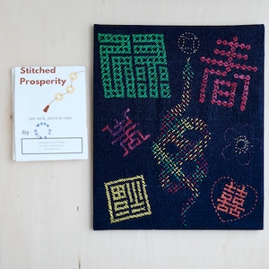 Puede incluir: Un panel de tela azul oscuro con bordados coloridos. El bordado incluye formas geométricas, flores y caracteres japoneses. El panel de tela está titulado "Stitched Prosperity" e incluye el texto "just stick, stitch & rinse".