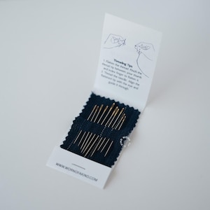 Könnte beinhalten: Ein weißes Heft mit Nähnadeln und Einfädel-Tipps. Das Heft ist geöffnet und zeigt einen dunkelblauen Stoff mit mehreren goldfarbenen Nadeln. Der Text "Threading Tips" und "WWW.WORNOFAFAKIND.COM" sind sichtbar.