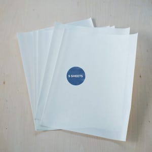 May include: A stack of five white paper sheets. A blue circular sticker on the top sheet reads "5 SHEETS". The paper has a slightly translucent quality, and the background is a light wood grain.