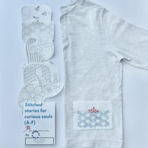 May include: A pale grey long-sleeved shirt with an embroidered pocket showing a red paper boat on blue waves. Next to the shirt are paper cutouts with stitched designs of boats, waves, and animals. A small book reads "Stitched stories for curious souls (A-F)".