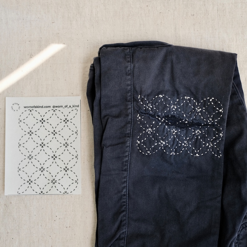 Puede incluir: Un par de pantalones vaqueros azul oscuro con un dise&ntilde;o bordado blanco en la rodilla. El dise&ntilde;o es un patr&oacute;n repetitivo de c&iacute;rculos y estrellas. Los vaqueros est&aacute;n doblados por la mitad y sobre una superficie beige claro. El texto "wornofakind.com @worn_of_a_kind" est&aacute; impreso en una tarjeta blanca junto a los vaqueros.