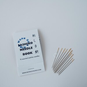 K&ouml;nnte beinhalten: Ein wei&szlig;es Nadelbuch mit blauem Text "Worn Sashiko Needle Book" und japanischen Schriftzeichen. Zehn verschiedene Sashiko-Nadeln sind neben dem Buch aufgef&auml;chert. Die Nadeln haben goldfarbene &Ouml;sen.