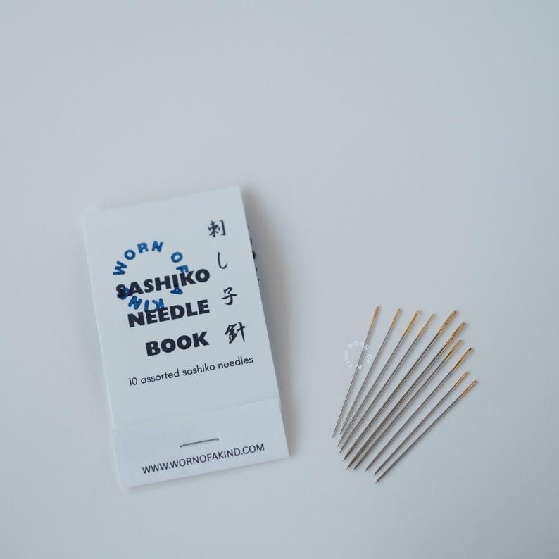 K&ouml;nnte beinhalten: Ein wei&szlig;es Nadelbuch mit blauem Text "Worn Sashiko Needle Book" und japanischen Schriftzeichen. Zehn verschiedene Sashiko-Nadeln sind neben dem Buch aufgef&auml;chert. Die Nadeln haben goldfarbene &Ouml;sen.