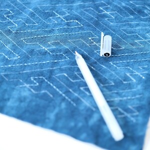May include: A white fabric with a blue dye pattern and a white fabric pen lying on top of it. The fabric has a stitched pattern of lines and squares.