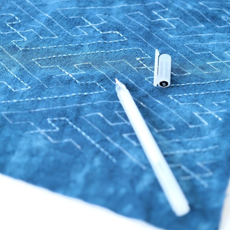 May include: A white fabric with a blue dye pattern and a white fabric pen lying on top of it. The fabric has a stitched pattern of lines and squares.