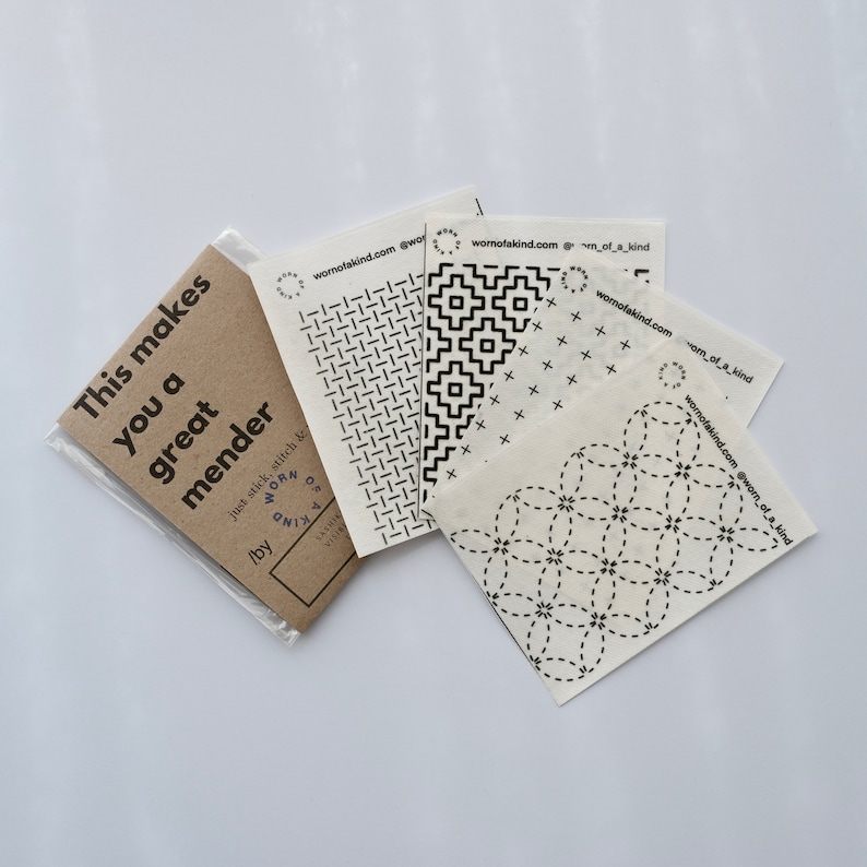 Puede incluir: Cuatro cuadrados de tela blanca con dise&ntilde;os de costura negros. Los cuadrados est&aacute;n etiquetados como "worn of a kind" y tienen la direcci&oacute;n del sitio web "wornofakind.com". Una tarjeta marr&oacute;n con el texto "This makes you a great mender" est&aacute; en el fondo.