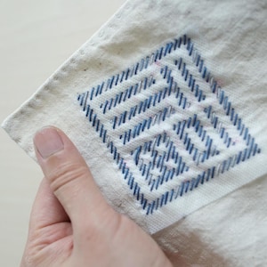 May include: Close-up of a square, off-white fabric patch with a geometric design embroidered in blue thread. The design features a series of parallel lines and angled segments, creating a maze-like pattern. The fabric is held by a hand.