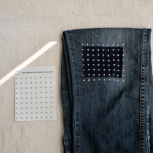 Puede incluir: Un par de pantalones vaqueros azules con un parche de punto de cruz blanco en la rodilla. El parche es un cuadrado con una cuadr&iacute;cula de cruces blancas sobre un fondo azul oscuro. Los vaqueros est&aacute;n colocados sobre una superficie beige claro. El texto "wornofakind.com @worn_of_a_kind" est&aacute; impreso en una tarjeta blanca a la izquierda de los vaqueros.
