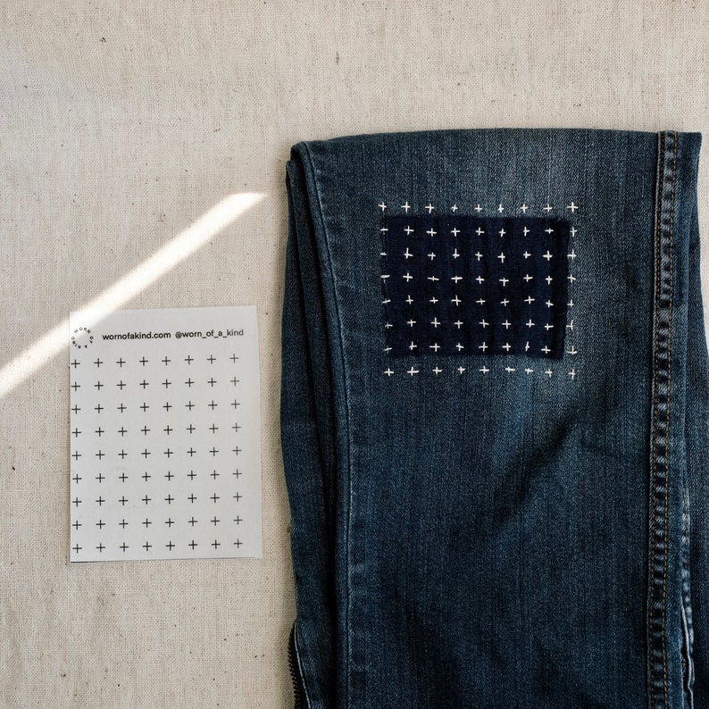 Puede incluir: Un par de pantalones vaqueros azules con un parche de punto de cruz blanco en la rodilla. El parche es un cuadrado con una cuadr&iacute;cula de cruces blancas sobre un fondo azul oscuro. Los vaqueros est&aacute;n colocados sobre una superficie beige claro. El texto "wornofakind.com @worn_of_a_kind" est&aacute; impreso en una tarjeta blanca a la izquierda de los vaqueros.