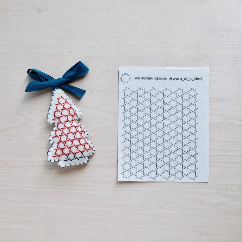 May include: A handmade Christmas tree ornament with red cross-stitch detailing on a white felt base. It has a dark blue ribbon for hanging. A white card with a geometric pattern and the text "wornofakind.com @worn_of_kind" is also present.