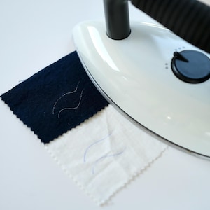 May include: A white handheld steamer with a black handle is shown steaming a dark blue fabric and a white fabric. Both fabrics have a wavy line drawn on them.