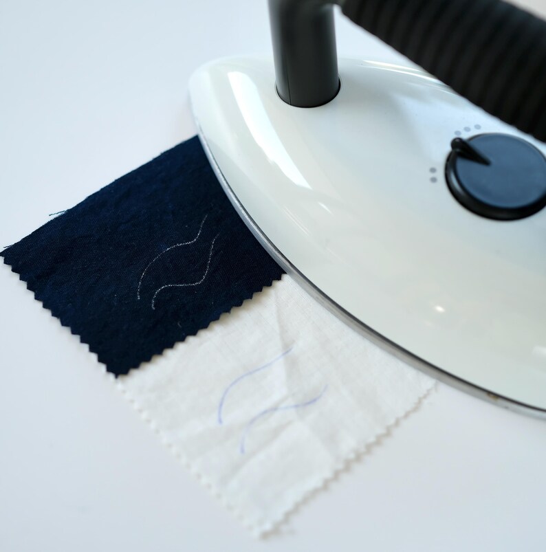 May include: A white handheld steamer with a black handle is shown steaming a dark blue fabric and a white fabric. Both fabrics have a wavy line drawn on them.