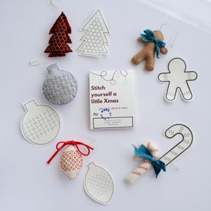 Könnte beinhalten: Eine Sammlung handgefertigter Weihnachtsschmuckstücke, darunter Filzbäume, Lebkuchenmänner und Zuckerstangen. Die Ornamente sind in verschiedenen Farben gehalten, mit roten, weißen und blauen Akzenten. Eine Karte mit dem Text "Stitch yourself a little Xmas" ist ebenfalls vorhanden.