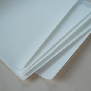 May include: A stack of white, textured paper sheets, fanned out to reveal the edges. The paper has a slightly off-white hue and a subtle, woven texture. The sheets are neatly stacked, suggesting a stationery or art supply product.