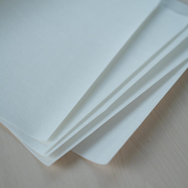 May include: A stack of white, textured paper sheets, fanned out to reveal the edges. The paper has a slightly off-white hue and a subtle, woven texture. The sheets are neatly stacked, suggesting a stationery or art supply product.