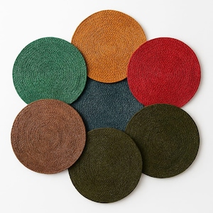May include: A collection of seven circular placemats arranged in a flower-like pattern. Each placemat features a tightly coiled, braided texture in distinct solid colors including mustard yellow, bright red, forest green, dark teal, brown, and olive.