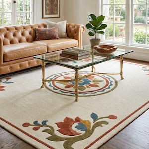 May include: A living room scene featuring a tufted leather sofa, a glass-top coffee table with gold legs, and an ornate rug with floral patterns. The rug has a cream background with red, blue, and green accents. A green armchair is on the right.