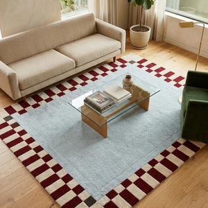 May include: A rectangular area rug featuring a large, solid light blue center panel surrounded by a wide border of alternating dark red and cream-colored squares. The rug has a textured, pile surface and is positioned on a light-colored wooden floor.