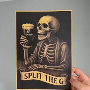 K&ouml;nnte beinhalten: Illustration eines Skeletts, das ein Pint Guinness h&auml;lt. Das Kunstwerk zeigt ein Skelett mit einem Bierglas. Der Text "SPLIT THE G" befindet sich auf einem Banner unter dem Skelett. Schwarzer Hintergrund.