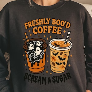 May include: Dark gray sweatshirt with the text "FRESHLY BOO'D COFFEE" in orange. The design features two coffee cups with Halloween-themed graphics, including ghosts, bats, and leaves. The phrase "SCREAM & SUGAR" is printed below the cups.