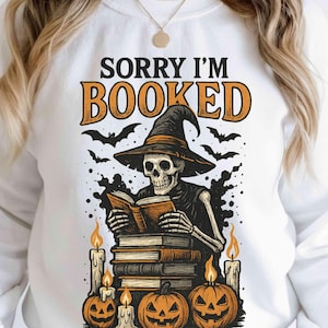 Peut inclure: Sweat-shirt blanc avec un motif sur le thème d'Halloween. Le motif représente un squelette lisant un livre, portant un chapeau de sorcière, entouré de citrouilles, de bougies et de chauves-souris. Le texte "SORRY I'M BOOKED" est au-dessus.