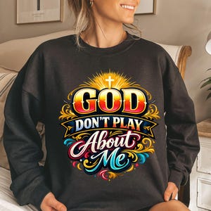 May include: Dark gray sweatshirt with a colorful graphic design. The design features the words "GOD DON'T PLAY ABOUT ME" in a stylized font, with a cross and sunburst design above the word "GOD".