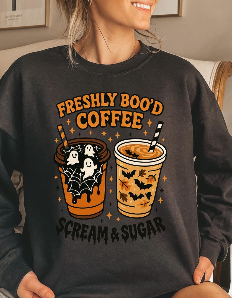May include: Dark gray sweatshirt with the text "FRESHLY BOO'D COFFEE" in orange. The design features two coffee cups with Halloween-themed graphics, including ghosts, bats, and leaves. The phrase "SCREAM & SUGAR" is printed below the cups.