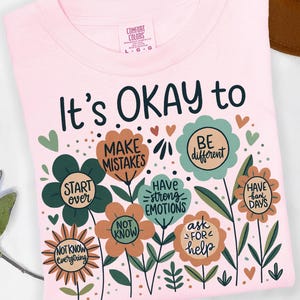 Op de afbeelding: Een lichtroze t-shirt met de tekst "It's OKAY to" boven kleurrijke bloemengraphics. Elke bloem heeft een andere boodschap, waaronder "Make Mistakes", "Be Different" en "Have Bad Days".