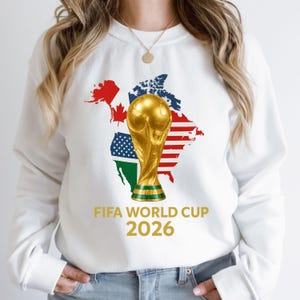 Puede incluir: Sudadera blanca con un gráfico dorado del trofeo de la Copa Mundial de la FIFA. El diseño incluye un mapa de América del Norte con las banderas de EE. UU., Canadá y México. El texto "FIFA WORLD CUP 2026" está impreso en dorado.