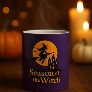 May include: A purple ceramic mug with a Halloween design. The mug displays a witch on a broomstick against a full moon, with the text "Season of the Witch". Steam rises from the mug, suggesting a hot drink.