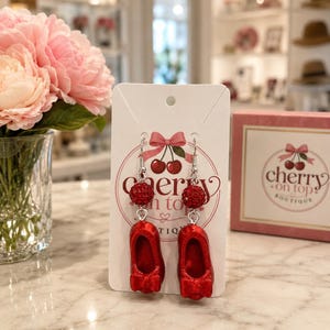 May include: A pair of dangle earrings on a white display card featuring the text Cherry on Top Boutique. Each earring consists of a silver-toned hook, a small red rhinestone-encrusted spherical bead, and a miniature red ballet flat shoe charm.