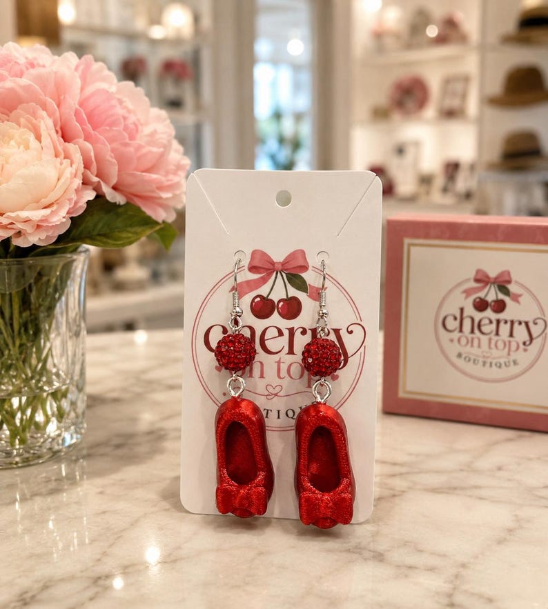 May include: A pair of dangle earrings on a white display card featuring the text Cherry on Top Boutique. Each earring consists of a silver-toned hook, a small red rhinestone-encrusted spherical bead, and a miniature red ballet flat shoe charm.
