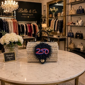 May include: A woven wicker handbag with a black handle and a decorative navy blue, red, and white embellishment. The handbag sits on a marble table in a boutique setting. The boutique's name, "Belle & Co. Boutique", is visible in the background.