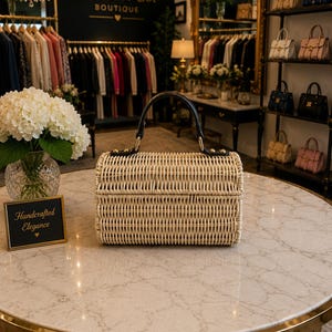 May include: A rectangular, woven rattan handbag with a black handle and gold-toned hardware sits on a marble table. The bag is light beige and has a structured design. The background shows a boutique setting with clothing racks and other handbags.