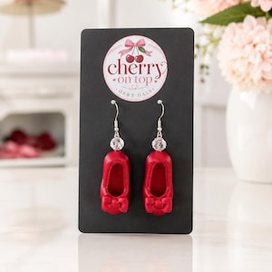 May include: A pair of dangle earrings featuring miniature red ballet slippers with small bows, accented by clear faceted beads on silver-toned hooks. The earrings are displayed on a black card with a logo reading cherry on top OERY HATS.