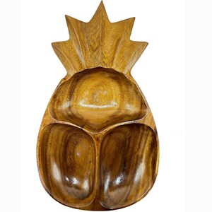 Puede incluir: Un plato de servicio de madera con forma de piña que presenta un acabado de veta de madera marrón natural. El plato está dividido en tres secciones distintas, con un compartimento superior más grande y dos inferiores más pequeños.