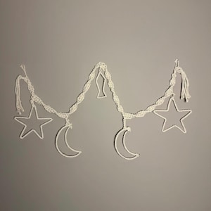 Peut inclure: Une guirlande en macramé blanc avec des formes d'étoiles et de croissants de lune, ainsi qu'un poisson décoratif. La guirlande est suspendue sur un fond gris, créant un thème céleste. Les étoiles et les lunes sont soulignées d'une texture tissée délicate.