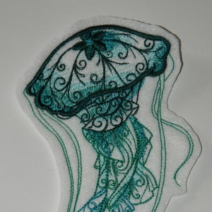 May include: A green and blue embroidered jellyfish with intricate details and flowing tentacles. The jellyfish is on a white background.