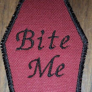 May include: A burgundy coffin-shaped patch with the words "Bite Me" embroidered in black script. The patch has a black stitched border. The background is a wooden surface.