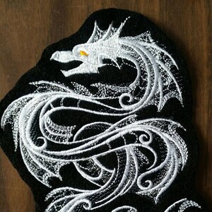 May include: White embroidered dragon design on a black fabric patch. The dragon is curled in a circle with its head facing the right side of the image. The dragon's eye is a bright yellow.