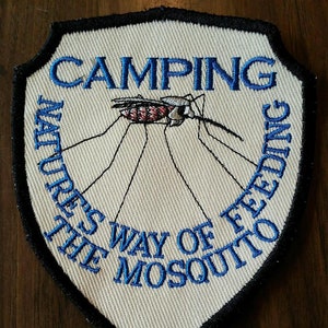 May include: A white embroidered patch with a black border in the shape of a shield. The patch features a blue inscription that reads "Camping Nature's Way of Feeding the Mosquito". A black and red mosquito is embroidered in the center of the patch.