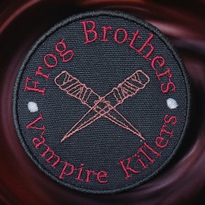 May include: A black circular patch with red embroidered text that reads "Frog Brothers Vampire Killers". The patch features two crossed daggers with a detailed design.