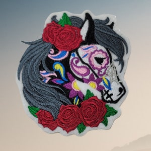 May include: Embroidered iron-on patch featuring a black and white horse with a sugar skull design. The horse has red, pink, blue, yellow, and purple details. The horse is surrounded by red roses with green leaves.
