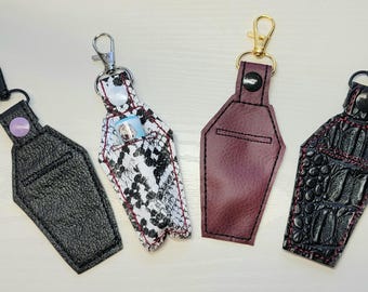 May include: Four different styles of black, white and burgundy faux leather coffin-shaped keychains with silver and gold metal clasps. Each keychain has a different pattern and design.