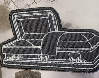 Pode incluir: Um remendo bordado preto que representa um caixão branco com contorno branco e tampa fechada. O caixão tem um contorno branco de uma alça de cada lado.