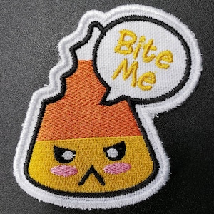 May include: Embroidered patch featuring a candy corn design with an angry face and pink blush. A speech bubble above the candy corn says "Bite Me" in yellow letters. The patch has a white border with a black outline.