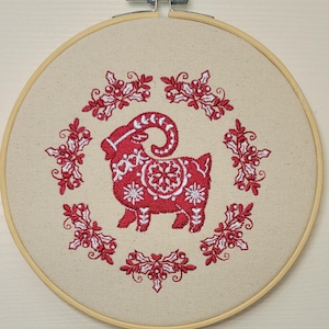 May include: A red and white embroidered goat design on a beige fabric stretched within a wooden embroidery hoop. The goat is surrounded by a wreath of red holly and white accents, creating a festive, handcrafted look.