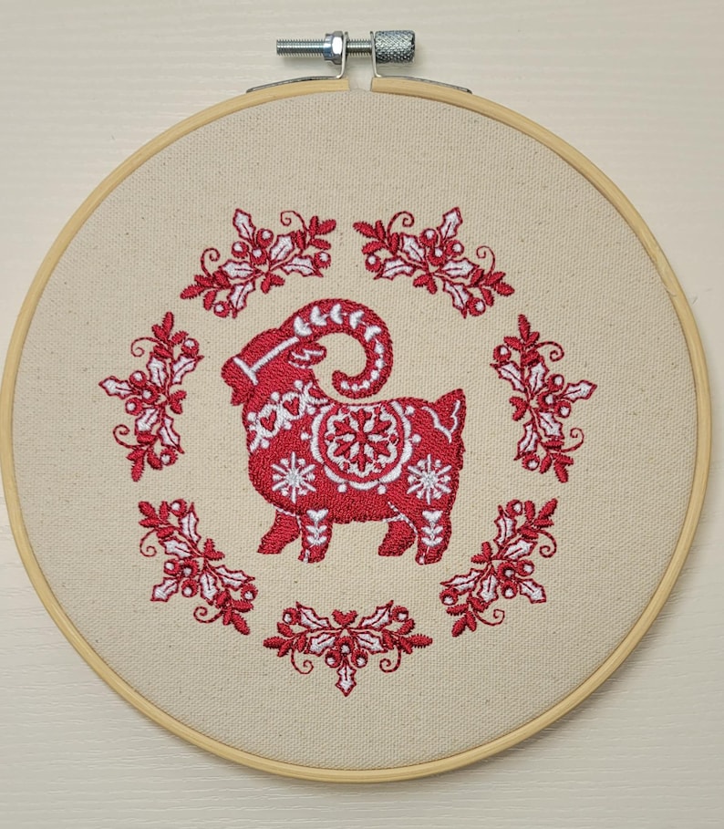 May include: A red and white embroidered goat design on a beige fabric stretched within a wooden embroidery hoop. The goat is surrounded by a wreath of red holly and white accents, creating a festive, handcrafted look.