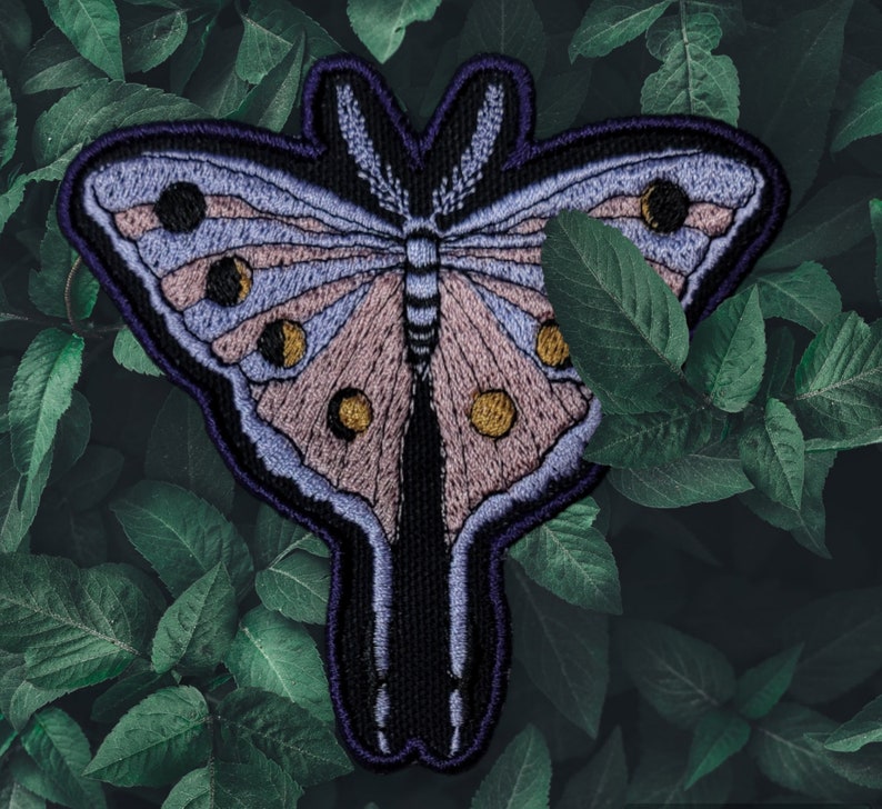 Op de afbeelding: Geborduurde vlinderpatch met een gedetailleerd ontwerp. De patch heeft een mot met lichtpaarse en bruine vleugels, geaccentueerd met gele cirkels. De patch is omlijnd in donkerpaars en zwart, tegen een achtergrond van groene bladeren.